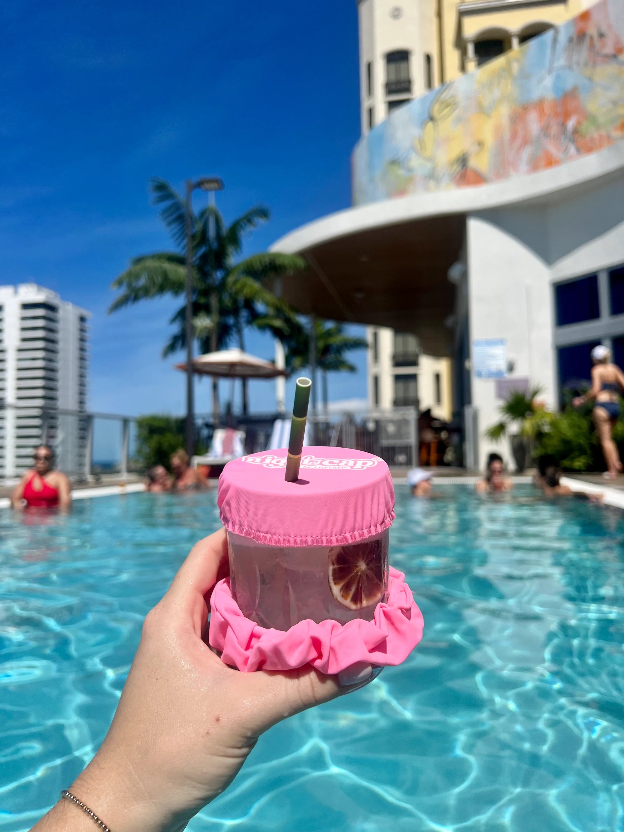 Pink NightCap drink cover protecting a cocktail with a straw while relaxing in a rooftop swimming pool.
